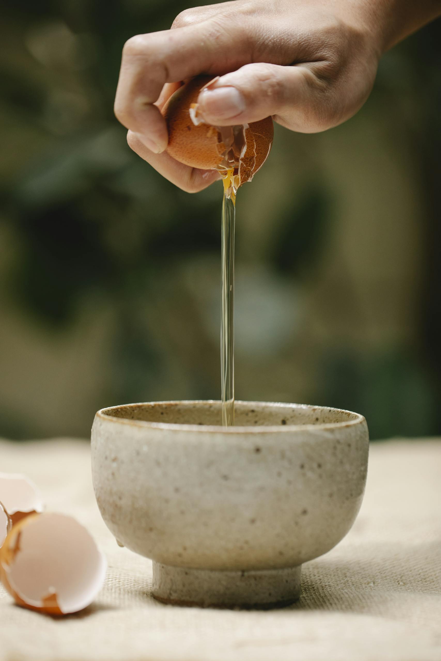 Crop anonymous female adding egg white from broken chicken egg into ceramic bowl on table