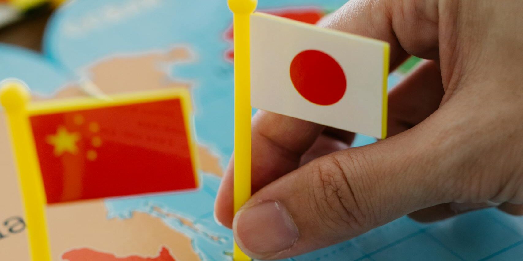 A hand holding the Japanese flag pinpoints Japan on a world map alongside China and India flags.
